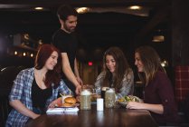 Glückliche Freundinnen genießen Essen in der Bar, während der Kellner Essen serviert — Stockfoto