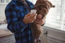 Vater benutzt Handy, während er sein Baby in der Küche hält — Stockfoto
