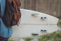 Mid section of a man with backpack carrying a surfboard walking through trail — Stock Photo