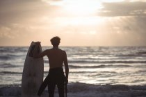 Rückansicht eines Mannes mit Surfbrett, der in der Abenddämmerung am Strand steht — Stockfoto