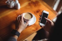 Mujer usando el teléfono móvil mientras toma una taza de café en la cafetería - foto de stock