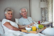 Couple aîné tenant le plateau de petit déjeuner sur le lit dans la chambre — Photo de stock