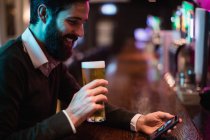 Homem olhando para o telefone celular enquanto toma um copo de cerveja no balcão do bar — Fotografia de Stock