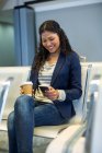Пригородная женщина с кофейной чашкой с помощью мобильного телефона в зоне ожидания в терминале аэропорта — стоковое фото