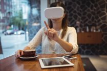 Бизнесвумен, сидя за столиком кафе с кофе, планшетом и телефоном, пользуется гарнитурой виртуальной реальности — стоковое фото