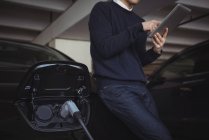 Man using digital tablet while charging electric car in garage — Stock Photo