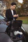 Человек, использующий мобильный телефон при зарядке электромобиля на улице — стоковое фото