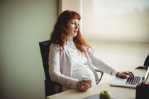 Donna d'affari incinta che utilizza il computer portatile mentre prende il caffè in ufficio — Foto stock