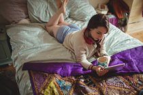 Belle femme utilisant une tablette numérique tout en prenant un café sur le lit à la maison — Photo de stock