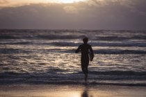 Silueta de un hombre llevando tabla de surf corriendo hacia el mar al atardecer - foto de stock