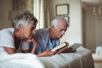 Femme âgée lisant un livre et homme âgé regardant tablette numérique sur le lit dans la chambre à coucher — Photo de stock