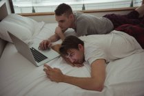 Гей-пара с помощью мобильного телефона и ноутбука на кровати в спальне — стоковое фото