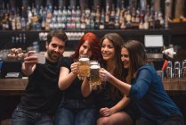 Amigos sosteniendo vasos de cerveza y tomando una selfie en el mostrador del bar usando el teléfono móvil - foto de stock