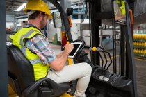 Travailleur masculin confiant utilisant une tablette numérique assis sur un chariot élévateur dans un entrepôt — Photo de stock