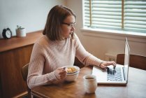 Mulher usando laptop enquanto toma café da manhã na sala de estar em casa — Fotografia de Stock