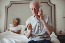 Preoccupato uomo anziano seduto in camera da letto con in mano la medicina e parlando sul telefono cellulare — Foto stock