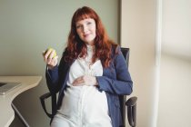 Портрет беременной предпринимательницы, держащей яблочные фрукты в офисе — стоковое фото
