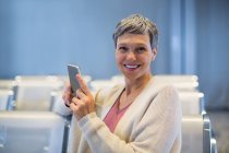 Портрет улыбающейся женщины, сидящей с мобильным телефоном в зале ожидания терминала аэропорта — стоковое фото
