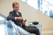 Retrato del hombre de negocios sonriente sentado con teléfono móvil en la zona de espera - foto de stock