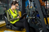 Trabalhadora confiante usando tablet digital enquanto se senta em empilhadeira no armazém — Fotografia de Stock