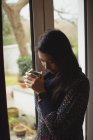 Задумчивая женщина пьет кофе у окна дома — стоковое фото