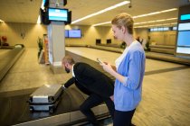 Man picking up suitcase on luggage conveyor belt while woman using mobile phone at airport terminal — Stock Photo
