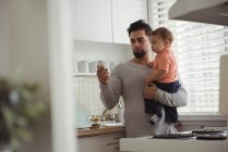 Vater benutzt Handy, während er Baby zu Hause in der Küche hält — Stockfoto