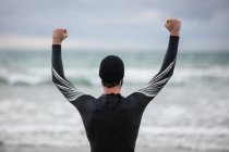 Vista traseira do atleta em terno molhado de pé com os braços para cima na praia — Fotografia de Stock