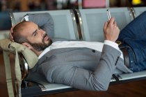 Бізнесмен використовує мобільний телефон, лежачи на стільцях в зоні очікування в терміналі аеропорту — стокове фото