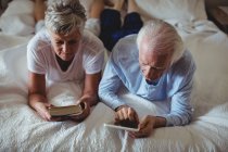 Старша пара використовує цифровий планшет і читає книгу на ліжку в спальні — стокове фото