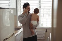 Father talking on mobile phone while holding baby in kitchen — Stock Photo