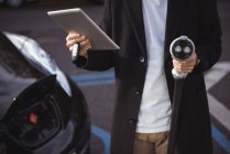 Sezione centrale dell'uomo che utilizza tablet digitale mentre tiene il caricabatterie per auto presso la stazione di ricarica del veicolo elettrico — Foto stock