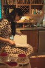 Mulher sentada na bancada da cozinha e livro de leitura em casa — Fotografia de Stock