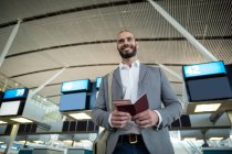 Hombre de negocios sonriente sosteniendo una tarjeta de embarque y pasaporte en la terminal del aeropuerto - foto de stock