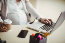 Partie médiane de la femme d'affaires enceinte utilisant un ordinateur portable tout en prenant un café au bureau — Photo de stock