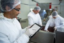 Carnicero usando tableta digital mientras sus compañeros trabajan en segundo plano en la fábrica de carne - foto de stock