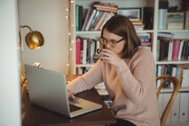 Mulher usando laptop enquanto bebe café na sala de estar em casa — Fotografia de Stock
