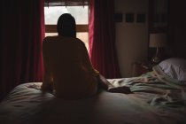 Жінка сидить на ліжку і дивиться через вікно вдома, вид ззаду — стокове фото
