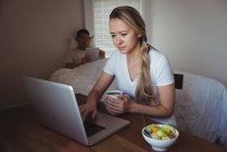 Жінка використовує ноутбук, маючи каву в спальні вдома — стокове фото