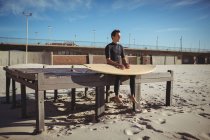 Задумчивый серфер сидит на деревянной платформе с доской для серфинга на пляже — стоковое фото