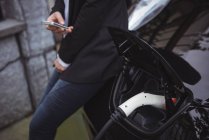 Seção média da mulher que usa o telefone móvel ao carregar o carro elétrico na estação de carregamento do veículo — Fotografia de Stock