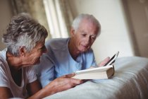 Старшая пара с помощью цифрового планшета и чтения книги на кровати в спальне — стоковое фото