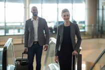 Smiling business people with luggage standing in front of an escalator at airport terminal — Stock Photo