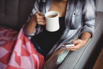 Женщина с помощью мобильного телефона, держа чашку кофе в гостиной дома — стоковое фото