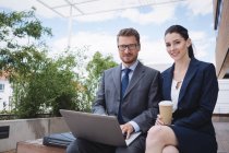 Porträt einer Geschäftsfrau und eines Kollegen, die vor einem Bürogebäude sitzen und Laptop benutzen — Stockfoto