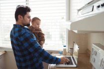 Отец использует ноутбук, держа ребенка на кухне дома — стоковое фото