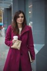 Femme d'affaires tenant tasse à café jetable, colis et journal tout en marchant sur le trottoir — Photo de stock