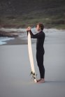 Вид збоку чоловіка в гідрокостюмі стоїть на пляжі з дошкою для серфінгу — стокове фото