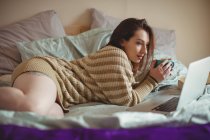 Bella donna che utilizza il computer portatile mentre prende il caffè sul letto a casa — Foto stock