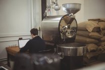 Man using laptop while sitting besides coffee roasting machine in coffee shop — Stock Photo